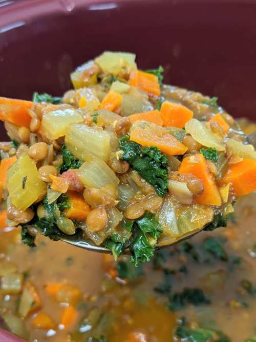A ladle holds a colorful mix of lentils, kale, carrots, and vegetables in a savory broth.
