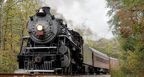 A vintage steam locomotive chugs along a tree-lined track, billowing white smoke into the air.