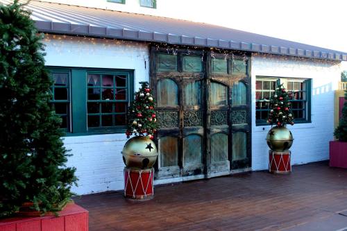 Decorative entrance with large wooden doors, flanked by Christmas trees and festive drum-shaped planters.