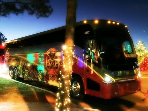 A brightly lit tour bus parked near a tree, with colorful lights reflecting off its surface at dusk.