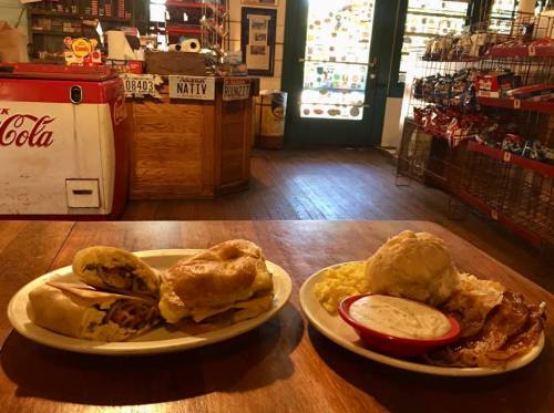 Two plates of food on a wooden table: one with sandwiches and the other with biscuits, gravy, eggs, and bacon.