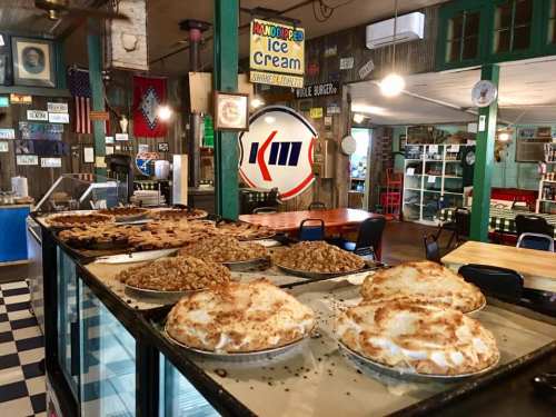 A cozy diner interior featuring a display of various pies on a counter, with vintage decor and seating in the background.