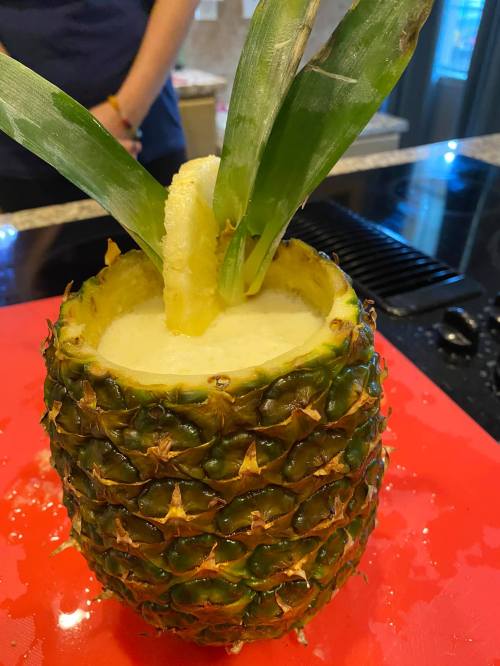 A fresh pineapple cut open at the top, filled with a creamy beverage and garnished with pineapple leaves.