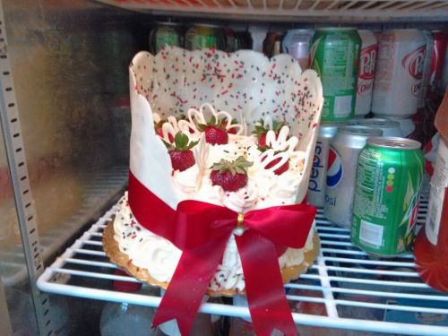 A decorated cake with strawberries and white frosting, topped with a red bow, inside a refrigerator among drinks.