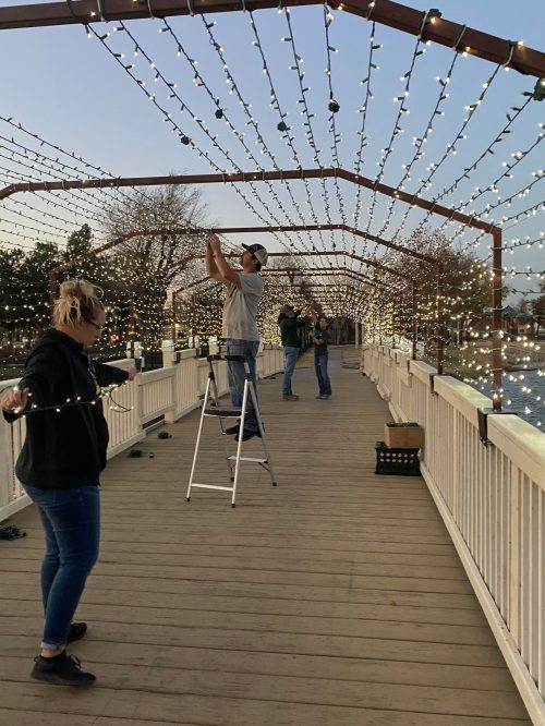 People decorating a bridge with string lights at dusk, with one person on a ladder and others assisting.