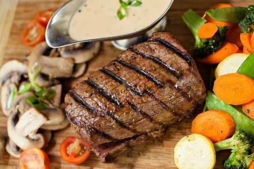Grilled steak with sauce, accompanied by sautéed vegetables and sliced mushrooms on a wooden cutting board.
