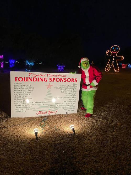 A person dressed as the Grinch stands beside a sign listing sponsors for a holiday event, with festive lights in the background.