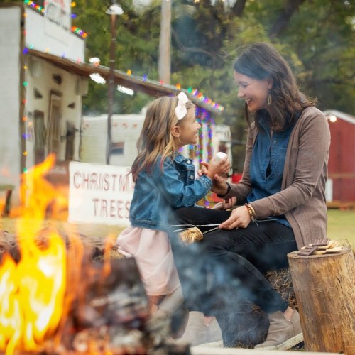 A woman and a girl sit by a campfire, smiling and enjoying each other's company in a festive outdoor setting.