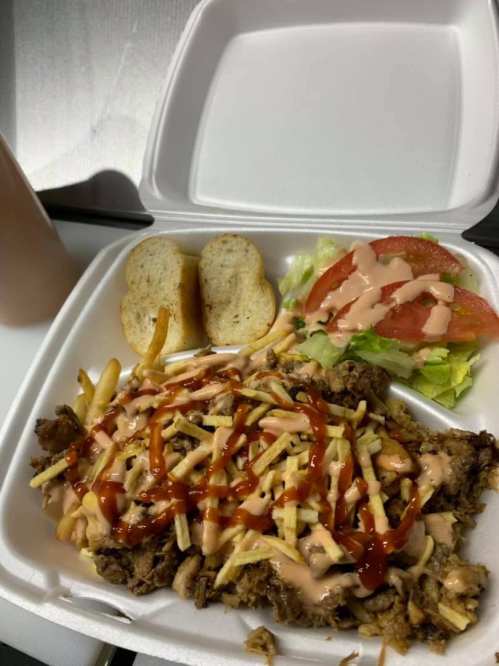 A takeout container with seasoned meat, fries, shredded cheese, lettuce, tomato, and sauce, alongside two slices of bread.