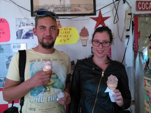 A man and a woman hold ice cream cones, smiling in a colorful shop decorated with photos and signs.