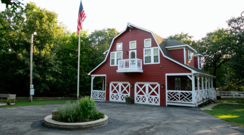 A red barn-style house with a porch, surrounded by trees and a flagpole, set in a peaceful outdoor area.