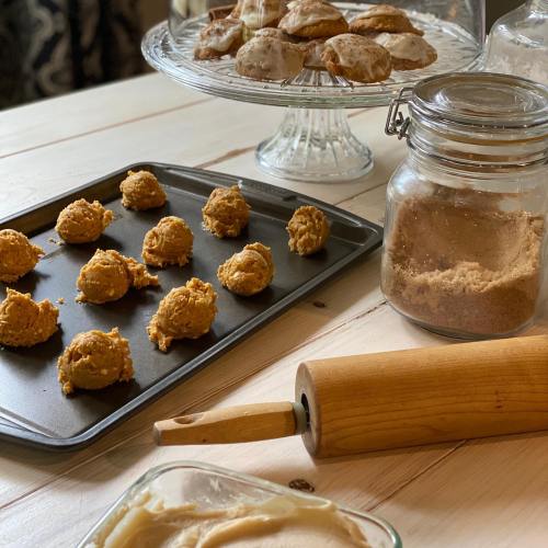 Baking scene with cookie dough on a tray, a jar of sugar, and baked cookies on a stand. Rolling pin in the foreground.