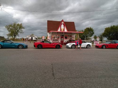 Two people stand between four colorful sports cars in front of a retro-style ice cream shop on a cloudy day.