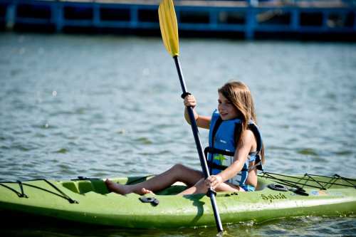 A young girl paddles a green kayak on a calm water body, wearing a life jacket and smiling.