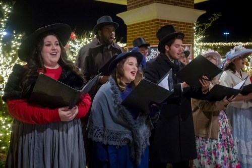 A group of carolers in festive attire sings outdoors, surrounded by twinkling lights and holiday decorations.