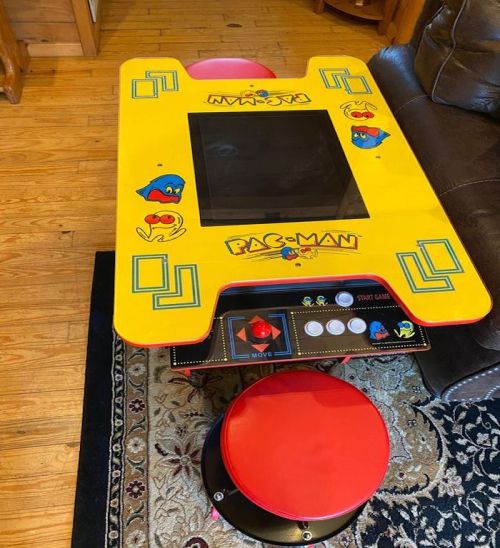 A colorful Pac-Man arcade table with a joystick and buttons, set on a patterned rug in a cozy room.