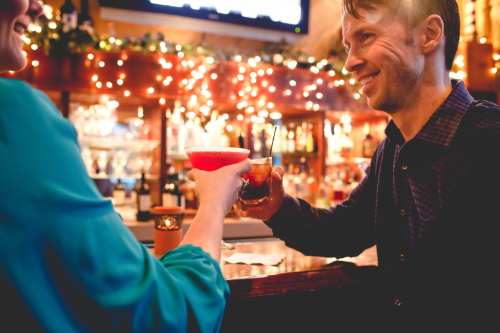 A man and woman clink glasses at a bar, surrounded by warm lights and a festive atmosphere.
