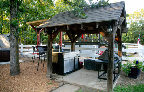 A rustic outdoor kitchen with a covered grill area, surrounded by trees and a patio with seating.