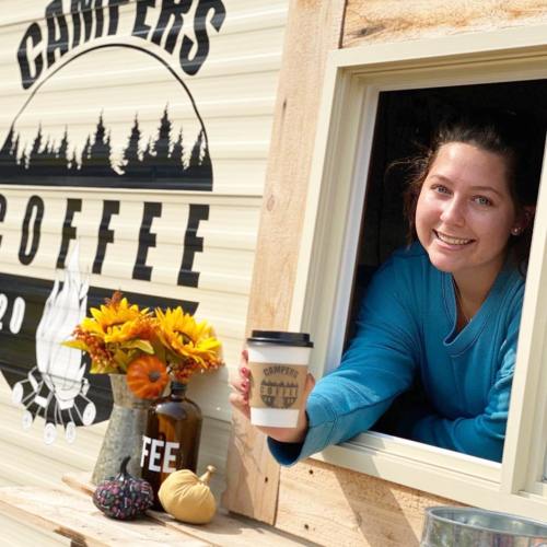 A smiling person in a blue sweater holds a coffee cup out of a window, with a camper-themed backdrop and autumn decor.
