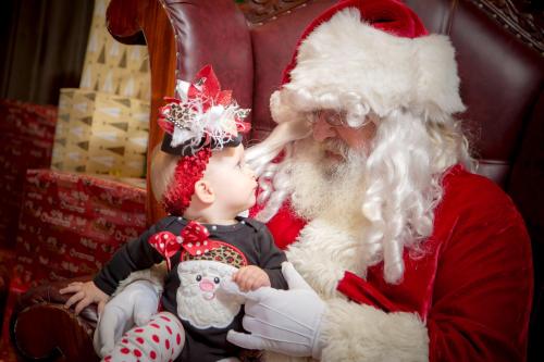 A baby in a festive outfit shares a sweet moment with Santa Claus, both smiling and looking at each other.