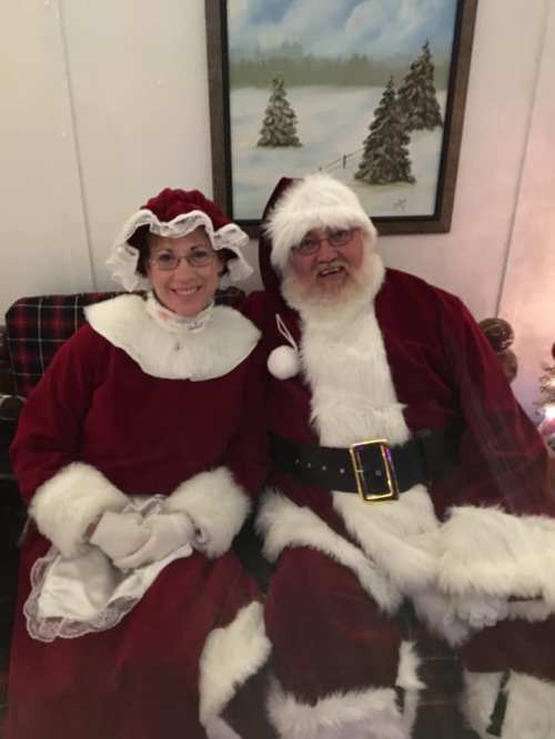 A smiling couple dressed as Santa and Mrs. Claus, sitting together in a cozy setting with holiday decorations.