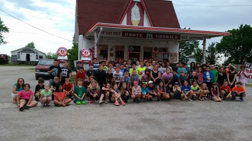 A large group of children and adults poses in front of a colorful ice cream shop on Route 66.