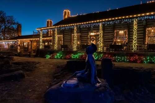 A log cabin adorned with colorful holiday lights at dusk, featuring a statue in the foreground.