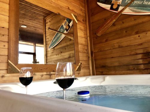 Two glasses of red wine sit on the edge of a hot tub, with a wooden wall and a mirror reflecting a surfboard in the background.