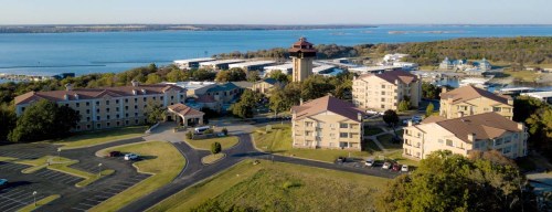 Aerial view of a lakeside resort with multiple buildings, a tower, and a marina in the background.