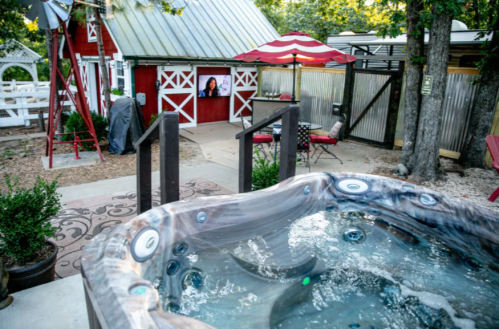 A hot tub in a backyard with a red barn, patio seating, and a TV screen in a cozy outdoor setting.