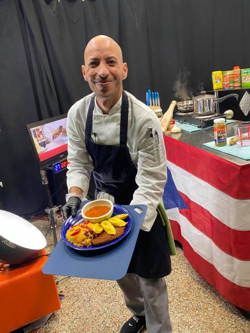 A chef in a kitchen holds a plate of food, smiling, with a Puerto Rican flag in the background.