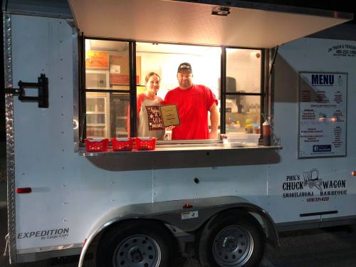 A food truck window with two people inside, holding a menu sign, ready to serve barbecue.