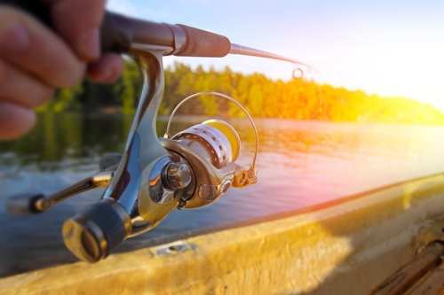 A close-up of a fishing rod and reel, with a scenic lake and sunset in the background.
