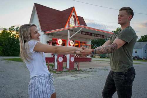 A couple joyfully holds hands and spins in front of a vintage gas station, with a sunset in the background.