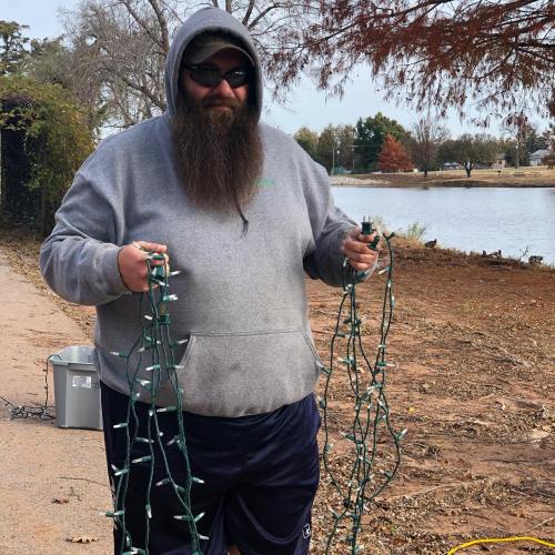 A man with a long beard wearing a hoodie holds two strands of holiday lights by a lake on a sunny day.