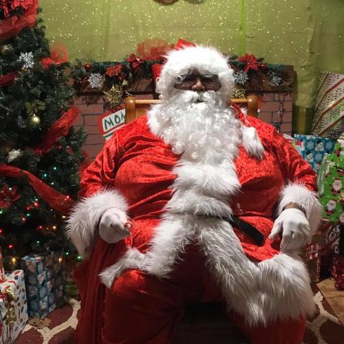A jolly Santa Claus in a red suit sits in front of a decorated Christmas tree and wrapped gifts.