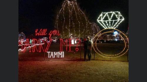 A couple stands beside a colorful light display that reads "Will you marry me?" with a diamond ring design.