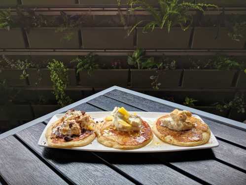 Three pancakes topped with whipped cream and syrup, served on a white plate against a backdrop of greenery.