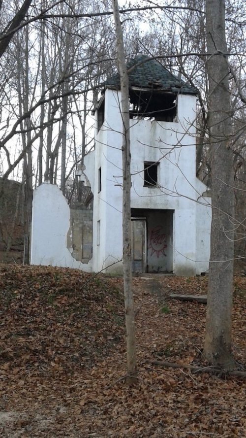 Abandoned white structure with a tower, surrounded by trees and fallen leaves in a wooded area.
