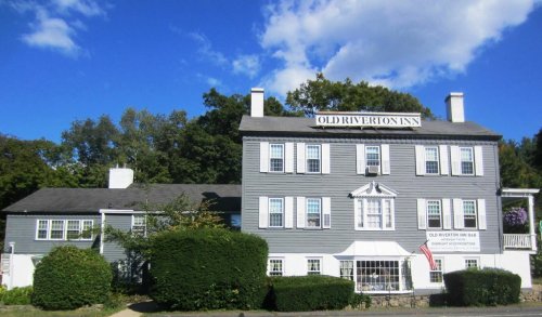 Historic Old Riverton Inn, a gray building with white trim, surrounded by greenery and under a blue sky with clouds.