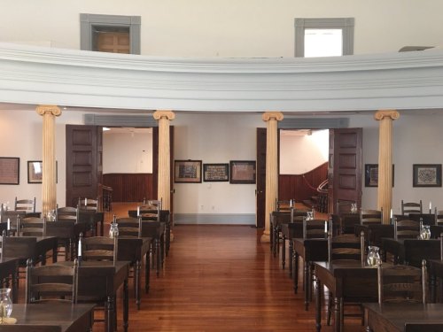 Interior of a historic room with wooden tables and chairs, featuring columns and framed documents on the walls.
