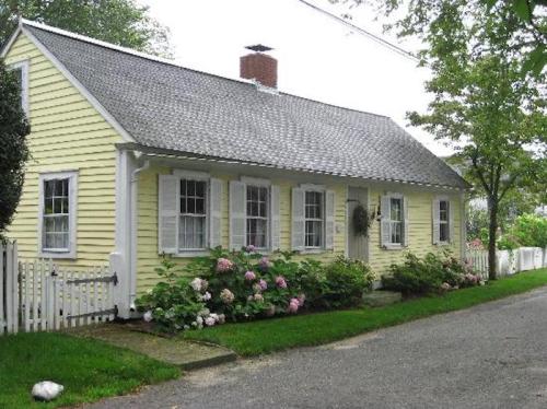 A charming yellow cottage with white trim, surrounded by blooming hydrangeas and a picket fence on a quiet street.