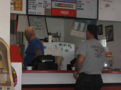 Two men stand at a counter in a casual eatery, with a menu and drink options visible in the background.