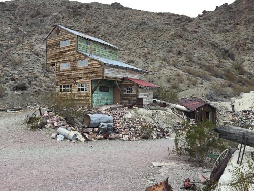 Abandoned wooden building in a desert landscape, surrounded by rocks and debris, with mountains in the background.