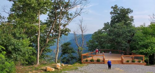 A scenic overlook with a wooden deck surrounded by trees, overlooking a mountainous landscape. People are enjoying the view.