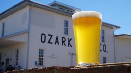 A glass of golden beer with a frothy head, set against a white building with "Ozark Co." painted on it.