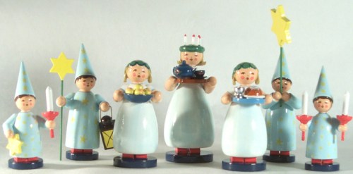 A group of colorful wooden figurines in festive attire, holding candles, stars, and various items, arranged in a cheerful display.