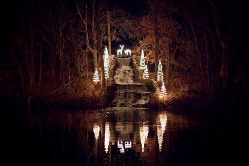 A waterfall illuminated by festive lights, surrounded by decorated trees, reflecting in the calm water at night.