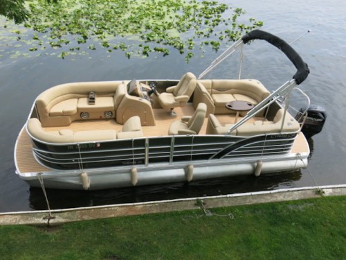 A spacious pontoon boat with beige seating and a canopy, docked by a calm lake surrounded by greenery.