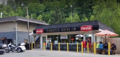 A drive-in restaurant with a Coca-Cola sign, featuring outdoor seating and a view of green trees in the background.